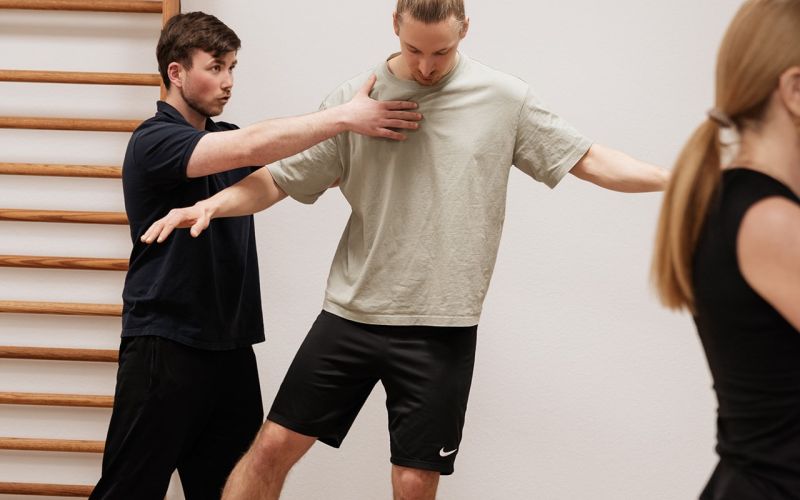Physiotherapeut unterstützt Mann beim Gleichgewichtstraining auf einem Balance-Kissen in der Praxis; im Hintergrund Sprossenwand, rechts unscharfe Person.