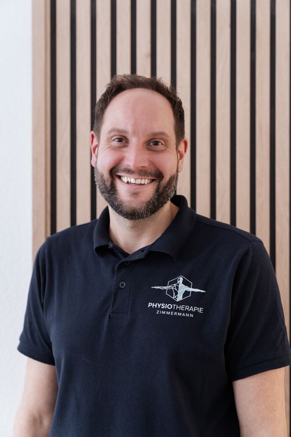 Porträt des lächelnden Physiotherapeuten Benjamin Zimmermann im Polo-Shirt „Physiotherapie Zimmermann“ vor einer Holzlamellenwand.