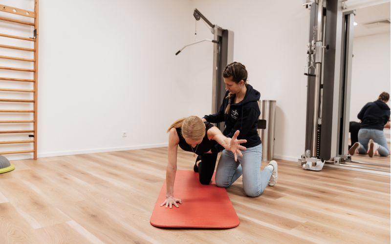 Physiotherapeutin begleitet eine Patientin bei einer Stabilisationsübung im Vierfüßlerstand auf einer Matte im Trainingsraum der Praxis.