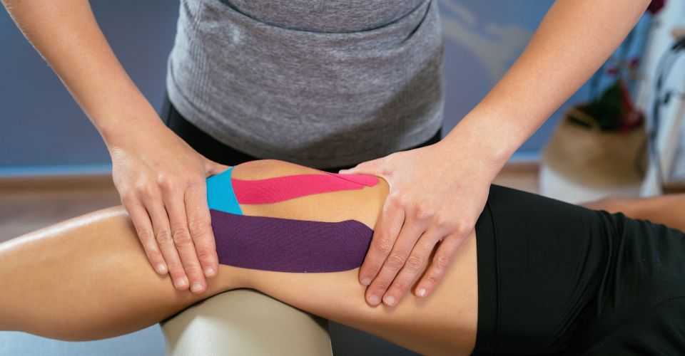 Nahaufnahme: Therapeutin legt bunte Kinesio-/Medical-Tapes auf den Oberschenkel einer Patientin zur Stabilisierung und Schmerzlinderung.