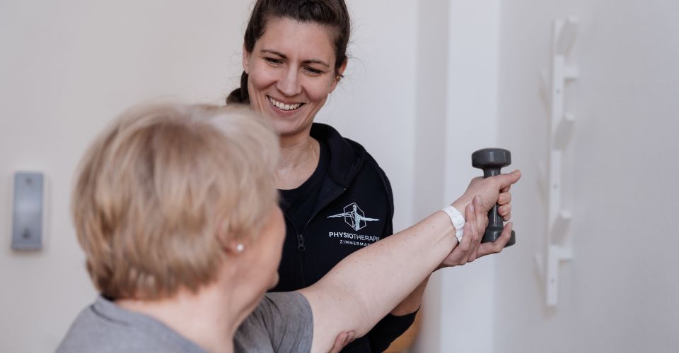 Physiotherapeutin unterstützt eine ältere Patientin bei einer Armübung mit Hantel zur Kräftigung und Mobilisation in der Praxis.