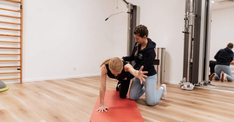 Patientin wird bei einer Übung auf der Bodenmatte von Physiotheapeutin gestützt 