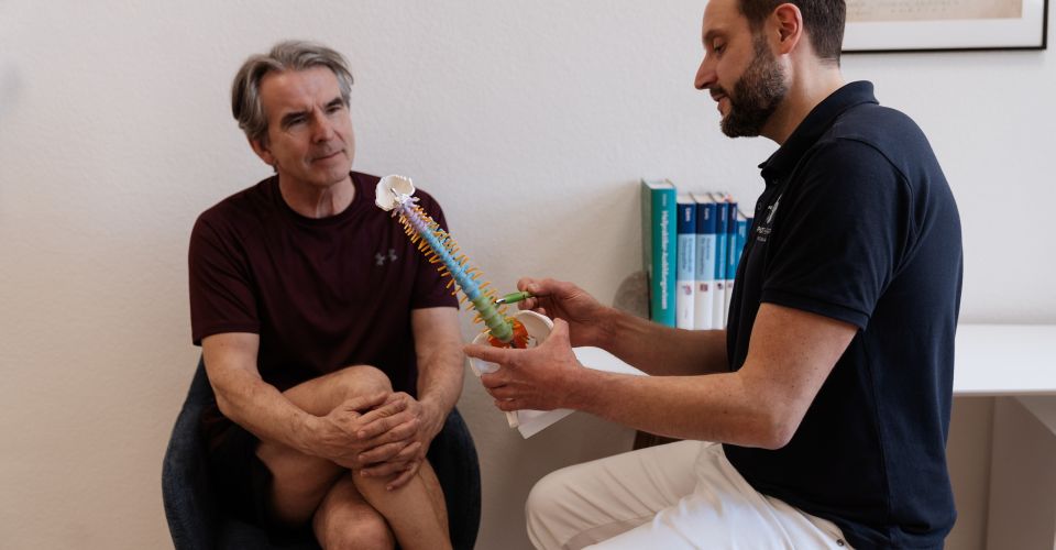 Physiotherapeut erklärt einem Patienten im Beratungsgespräch anhand eines Wirbelsäulenmodells die Behandlung in der Praxis.