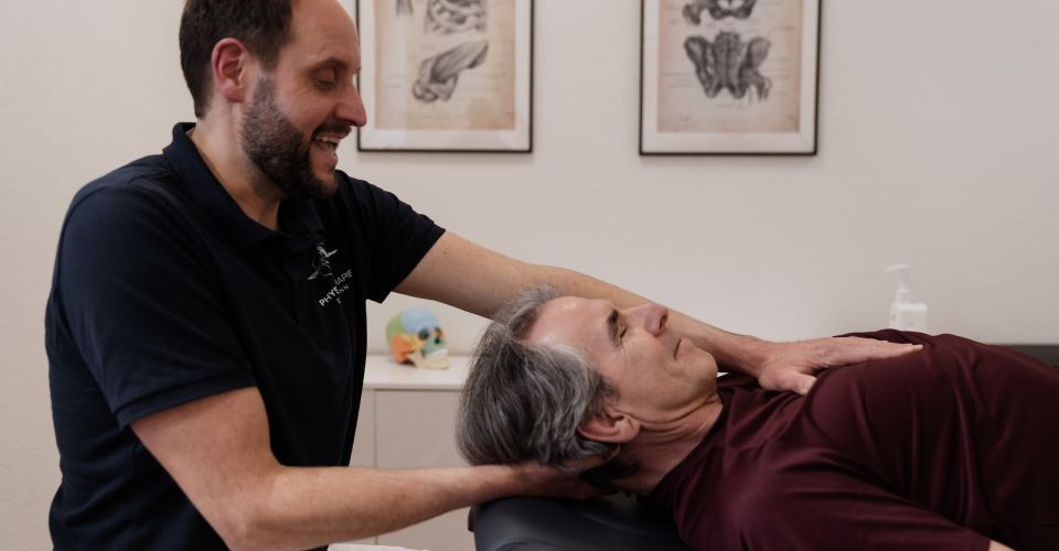 Osteopath führt parietale Behandlung im Nackenbereich eines Patienten auf der Therapieliege durch; im Hintergrund anatomische Poster.