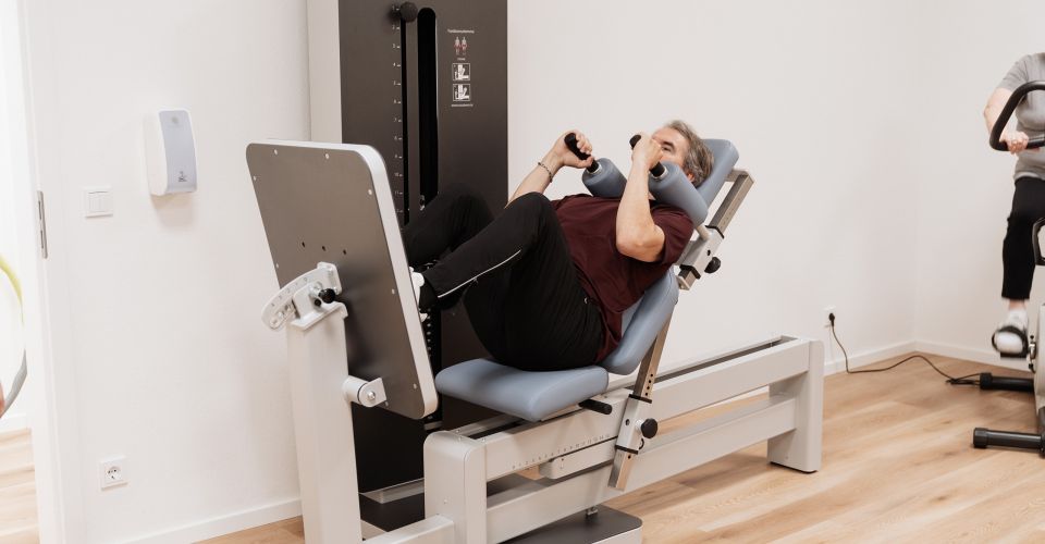Person trainiert in der Physiotherapiepraxis an einem Reha-Line-Beintrainer (Beinpresse) im Liegen; im Hintergrund weitere Trainingsgeräte und eine Person auf dem Ergometer.