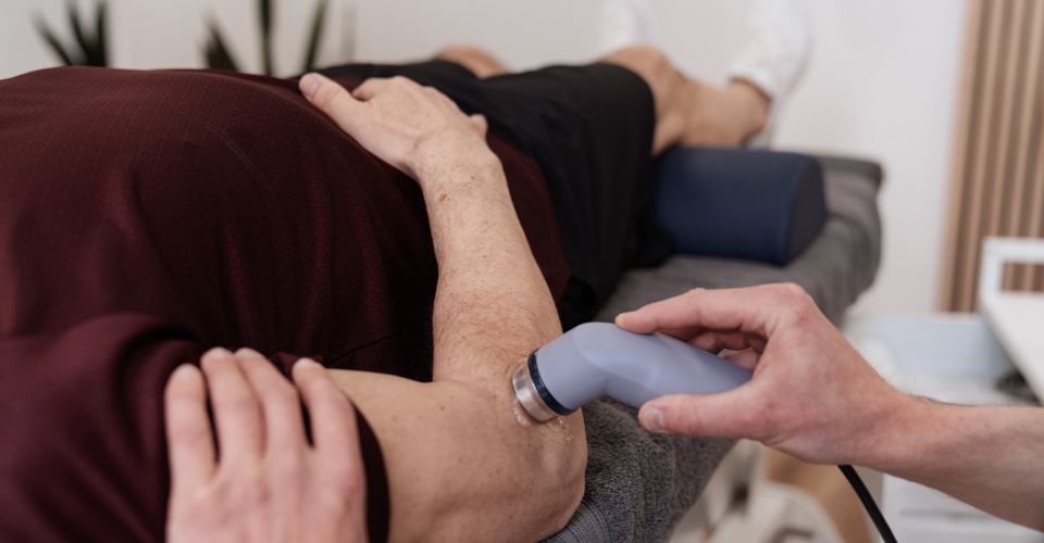 Patient liegt auf der Therapieliege, Therapeut behandelt den Ellenbogen mit einem Ultraschallgerät zur Schmerzlinderung und Regeneration.